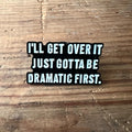 Nyssa pin with the phrase "I’ll get over it, just gotta be dramatic first" featuring bold lettering on a metal pin.