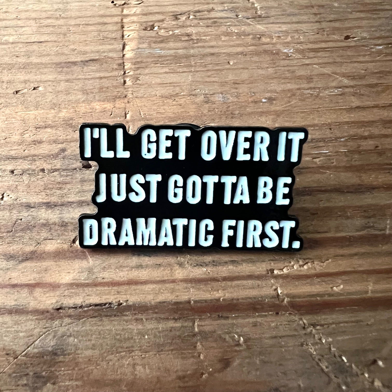 Nyssa pin with the phrase "I’ll get over it, just gotta be dramatic first" featuring bold lettering on a metal pin.