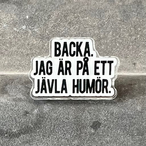 Colorful enamel pin with the phrase "Backa, jag är på ett jävla humör" from Nyssa, featuring bold text on a pin.