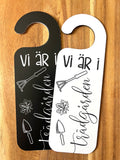 Nyssa Dörrhänge skylt with "Vi är i trädgården" text, hanging outdoors among garden plants and greenery.
